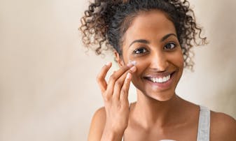 A woman smiles while putting lotion on her face.