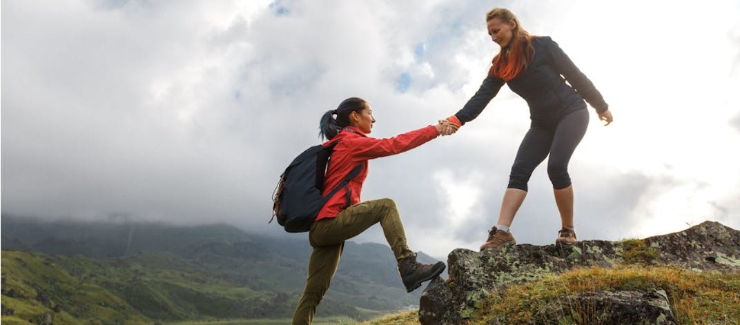 Two women helping each other climbing a mountain.