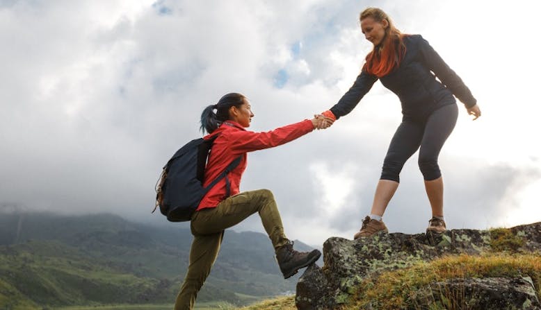Two women helping each other climbing a mountain.
