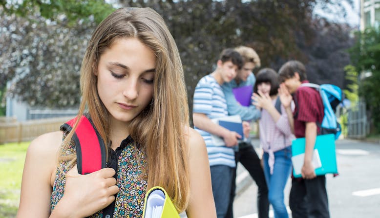 A teen girl looks sad in the foreground as a group of teenagers laugh behind their hands in the background.