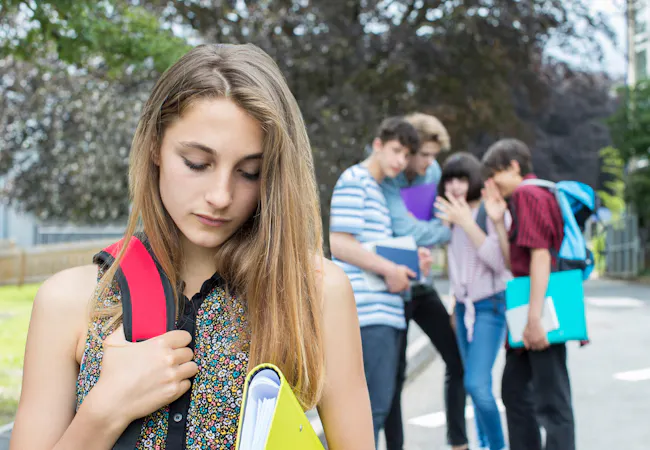 A teen girl looks sad in the foreground as a group of teenagers laugh behind their hands in the background.