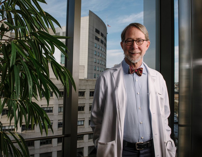 Philip Mease smiles in front of a large window while wearing a lab coat and bow tie. 