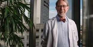 Philip Mease smiles in front of a large window while wearing a lab coat and bow tie. 