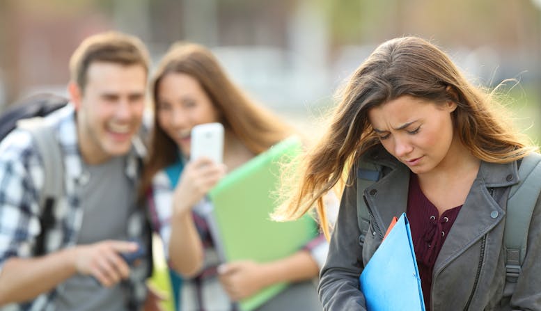 A teenage girl in the foreground looking upset while a teenage boy and girl in the background point and laugh while looking at a cell phone.