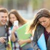 A teenage girl in the foreground looking upset while a teenage boy and girl in the background point and laugh while looking at a cell phone.