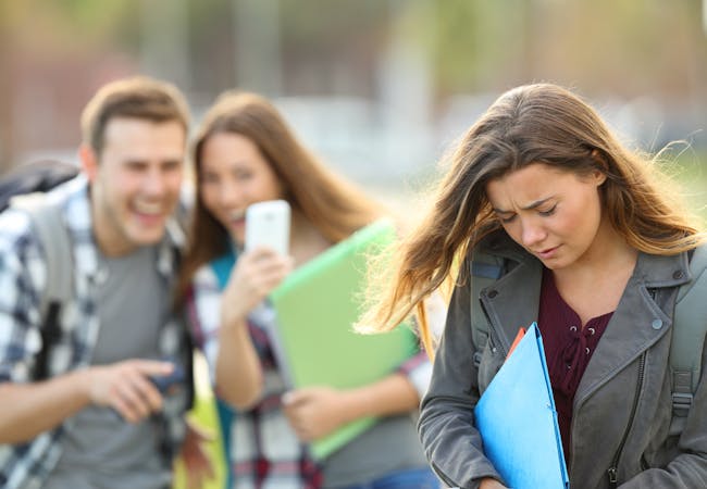 A teenage girl in the foreground looking upset while a teenage boy and girl in the background point and laugh while looking at a cell phone.