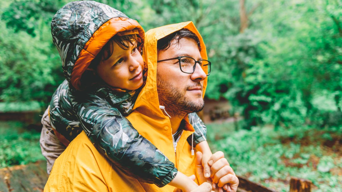 A father holds his son on his shoulders on a rainy day in the woods.