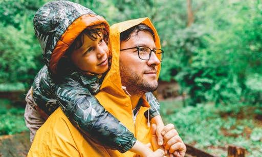 A father holds his son on his shoulders on a rainy day in the woods.