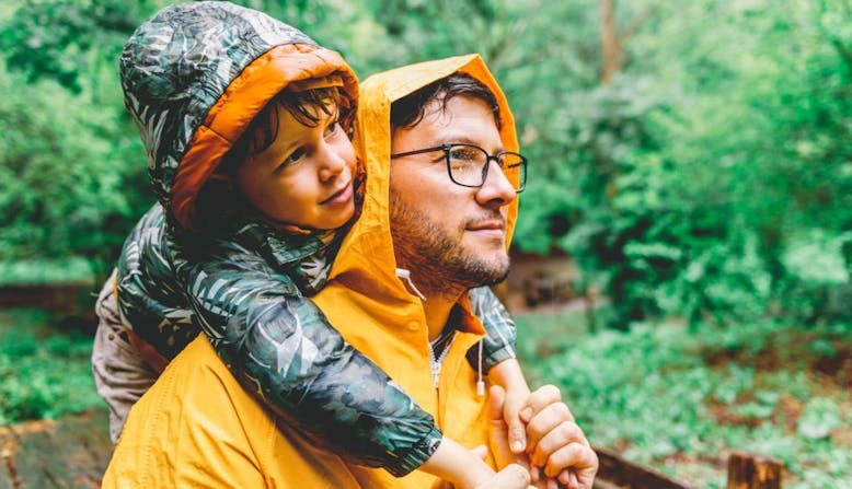 A father holds his son on his shoulders on a rainy day in the woods.