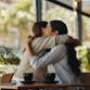 Two female friends sit together in a coffee shop while hugging and smiling.