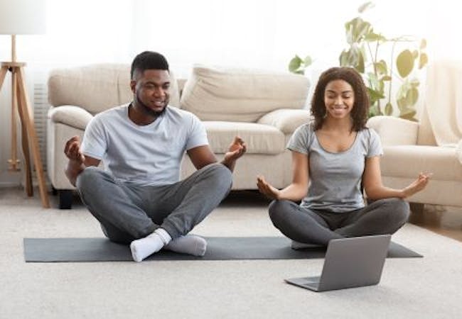 A man and woman meditating on a yoga mat while looking at a laptop.
