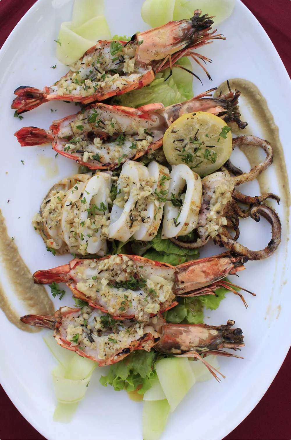 Grilled seafood platter featuring large prawns and whole squid seasoned with garlic herb butter, served on a white oval plate with fresh lettuce and lemon at a Bali seafood restaurant