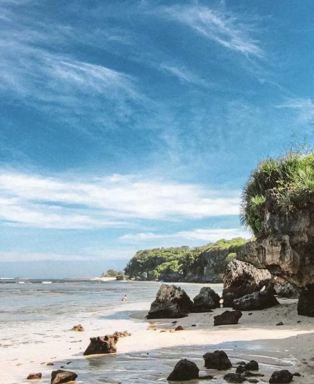 Scenic white sand beach at Nusa Dua, Bali, with volcanic rocks in the foreground, lush green cliffs, and calm turquoise waters under a bright blue sky