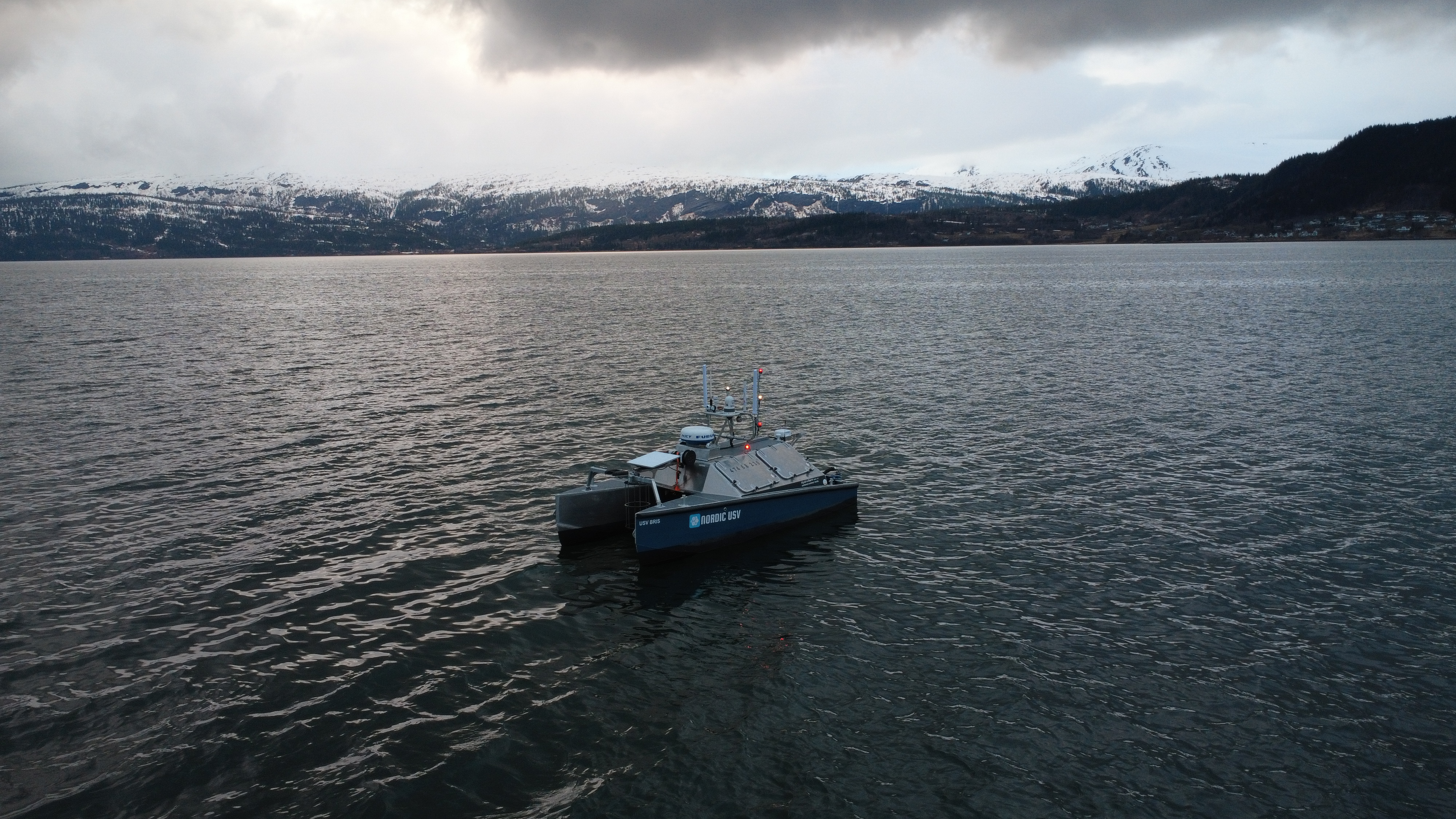 Ubemannet sjødrone med Nordic USV logo på rolig hav foran snødekte fjell. Dramatisk himmel med mørke skyer i skumringen.