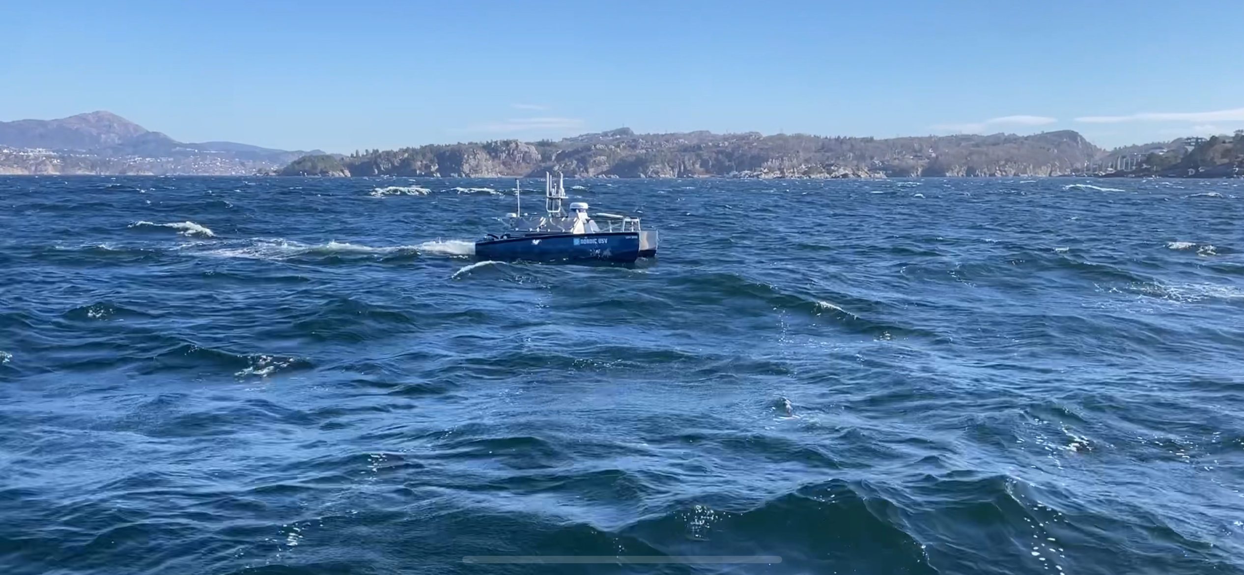 Blå og hvit ubemannet sjødrone med Nordic USV logo på åpent hav med bølger. Fjell og kystlinje i bakgrunnen under klar himmel.