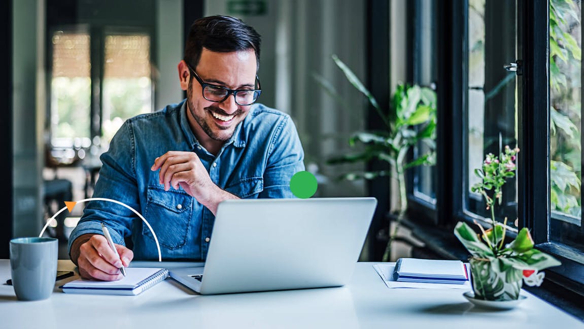 Man smiles while looking at laptop and writing in note pad.