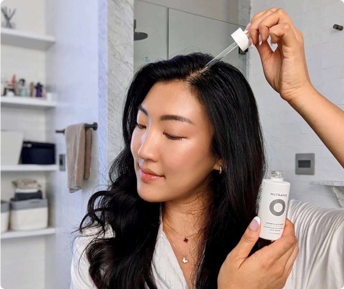 A woman applying Growth Activator Serum—a clear liquid in a dropper tip—to their part line.