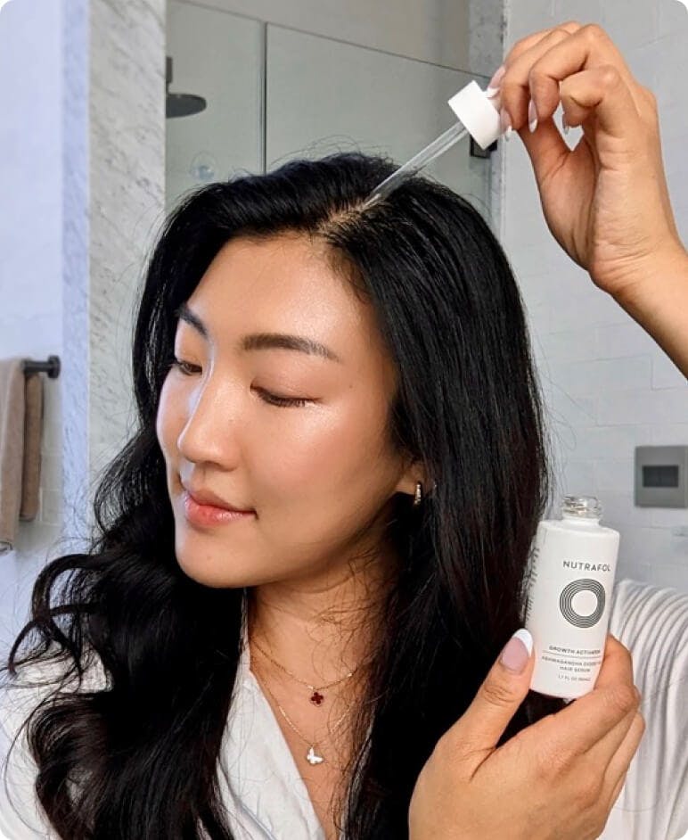 A woman applying Growth Activator Serum—a clear liquid in a dropper tip—to their part line.