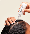 Hands applying Nutrafol Stress Reliever Essence—a clear liquid in a white bottle with nozzle—to the part line of their scalp.