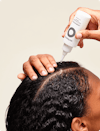 Hands applying Nutrafol Stress Reliever Essence—a clear liquid in a white bottle with nozzle—to the part line of their scalp.