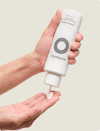 A hand holding a white bottle of Nutrafol Strand Defender Conditioner, dispensing a white cream into the other hand.