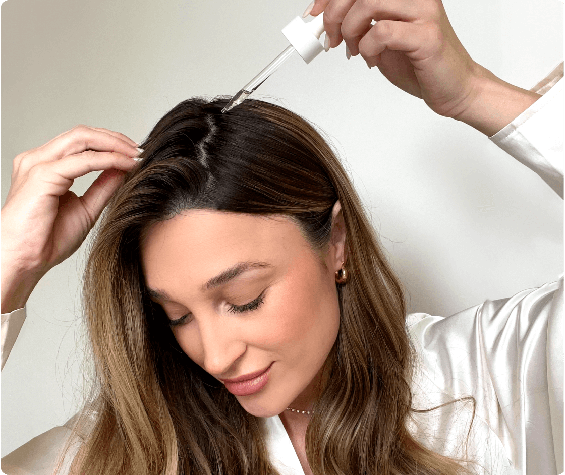 A woman applying Hair Serum—a clear liquid in a dropper tip—to their part line.