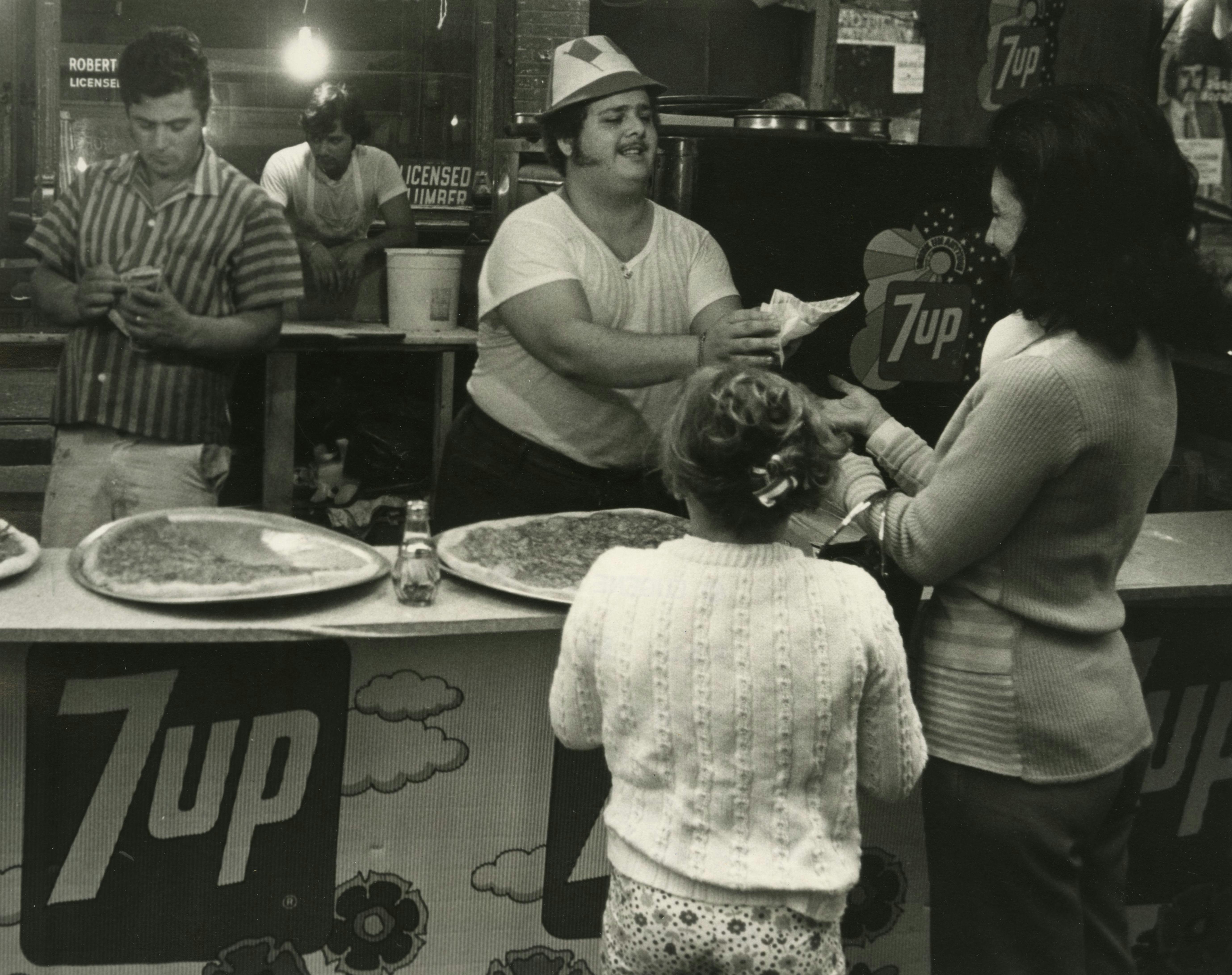 a black and white photo of a pizzeria and a counter man giving a woman and a young girl a slice