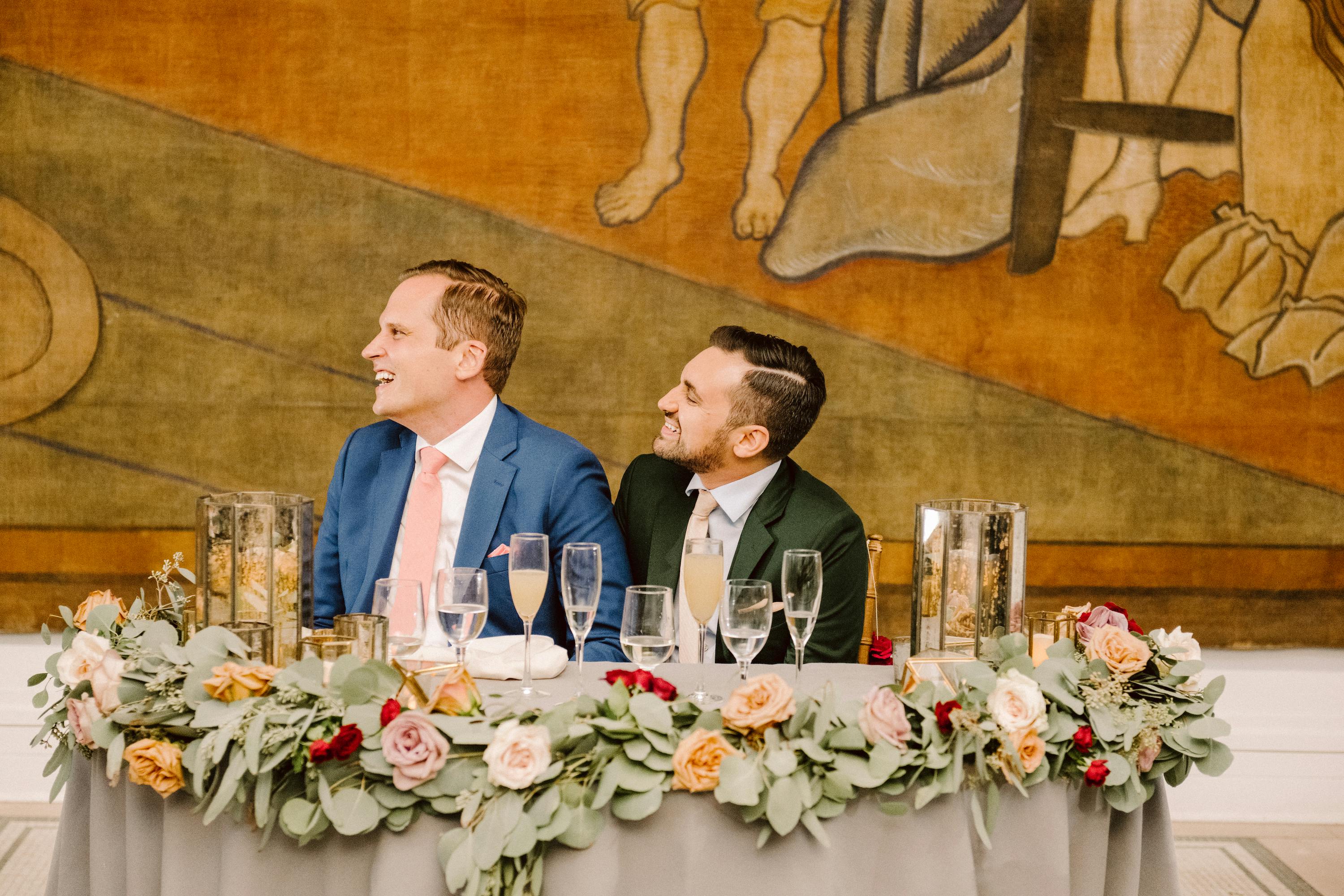 two men in suits sitting at a table during a celebration/wedding in Dexter gallery looking to the left and laughing