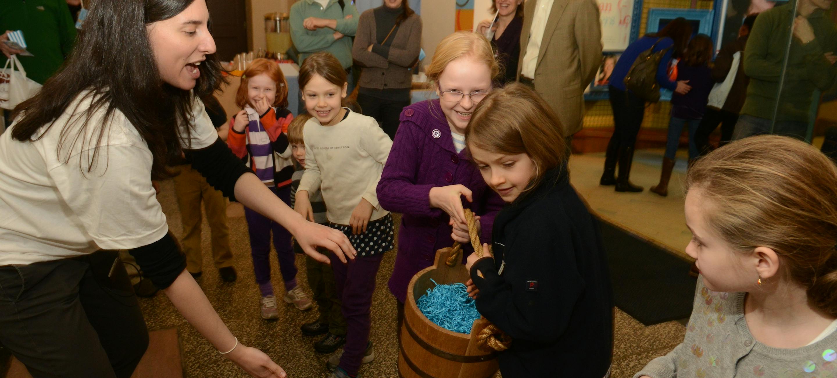 an educator working with a group of laughing kids to pass a historic water bucket filled with blue shredded paper
