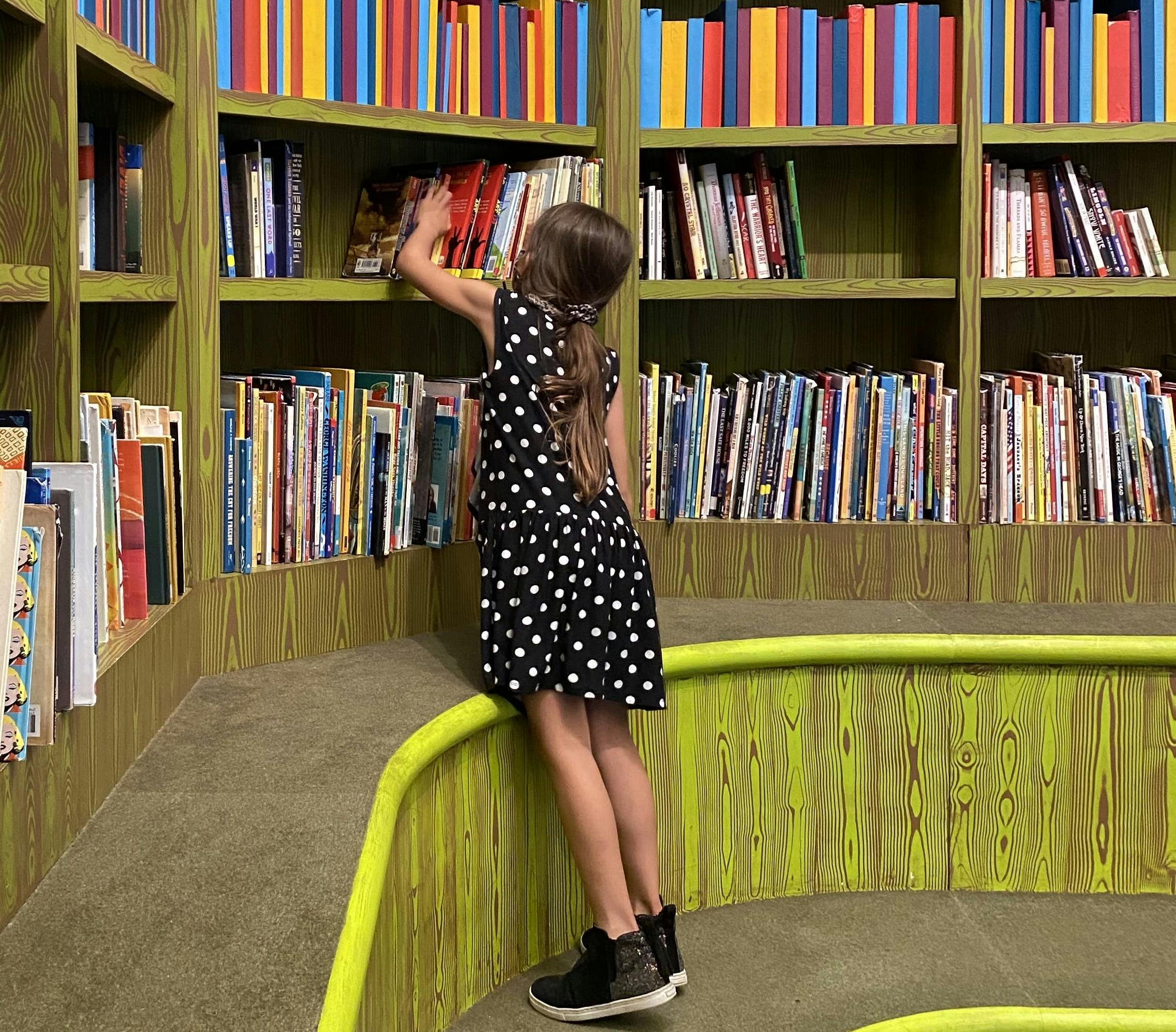 A young child faces away from the camera and reaches up to look at books in the barbara K. Lipman Library