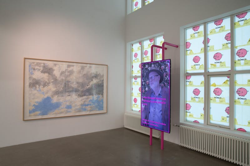 The picture shows two works of art. Left: On the wall hangs a large drawing by Geoffrey Hendricks, of a cloudy sky. Right: Video by Sæmundur Þór Helgason about poverty in Iceland. In the background, windows are covered with Bónus bags.