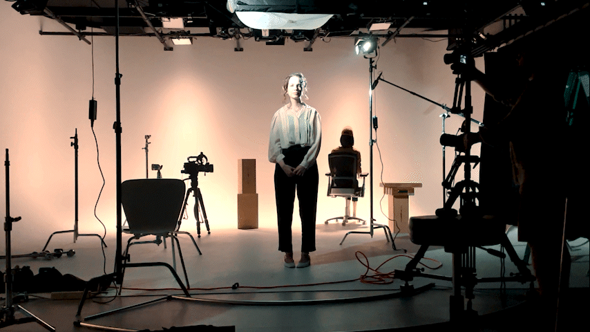 A woman stands in the middle of a film set as a man pushes a camera aimed at her on a dolly from right to left