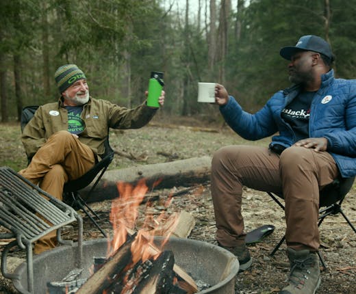 Two men cheers their drinks around a campfire.