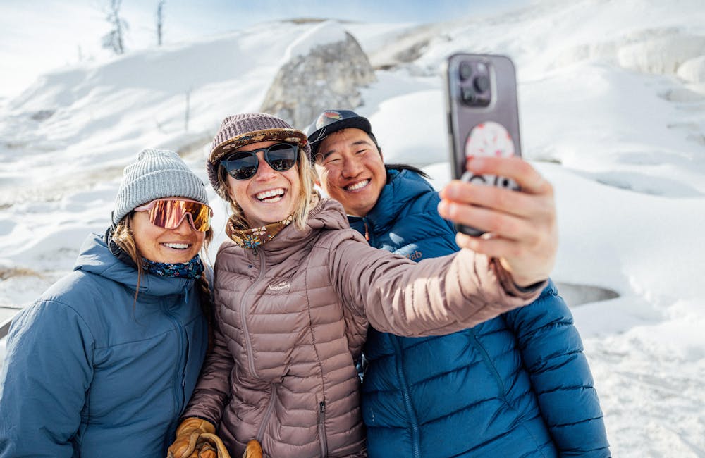 Three friends taking a selfie together at Yellowstone National Park in front snowy backdrop.