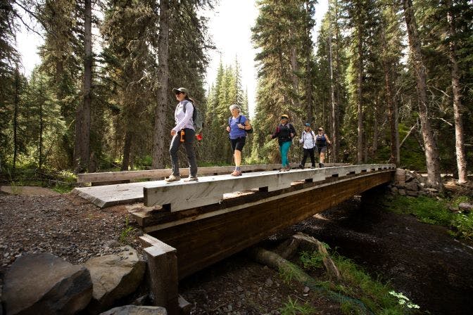 A group of women crosses a bridge across a creek while on a hiking trip wearing Oboz hiking shoes