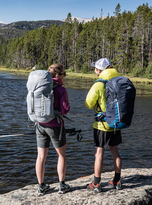 Two backpackers standing in front of a mountain lake wearing the Oboz Arete low hiking shoes.