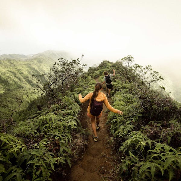 Two hikers navigating a muddy trail on a ridge of a tropical rainforest while wearing Oboz hiking shoes.