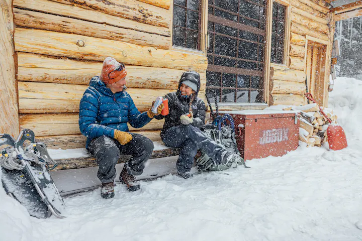 Two winter hikers smile and cheers beers while snowshoeing on a snowy day wearing Oboz Insulated hiking shoes