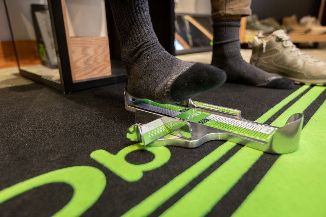 A person puts their foot on a Brannock Device branded with the Oboz logo.