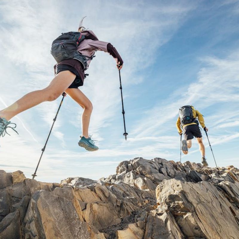 Two runners leap across scree terrain with treking poles in Oboz Katabatic shoes. 