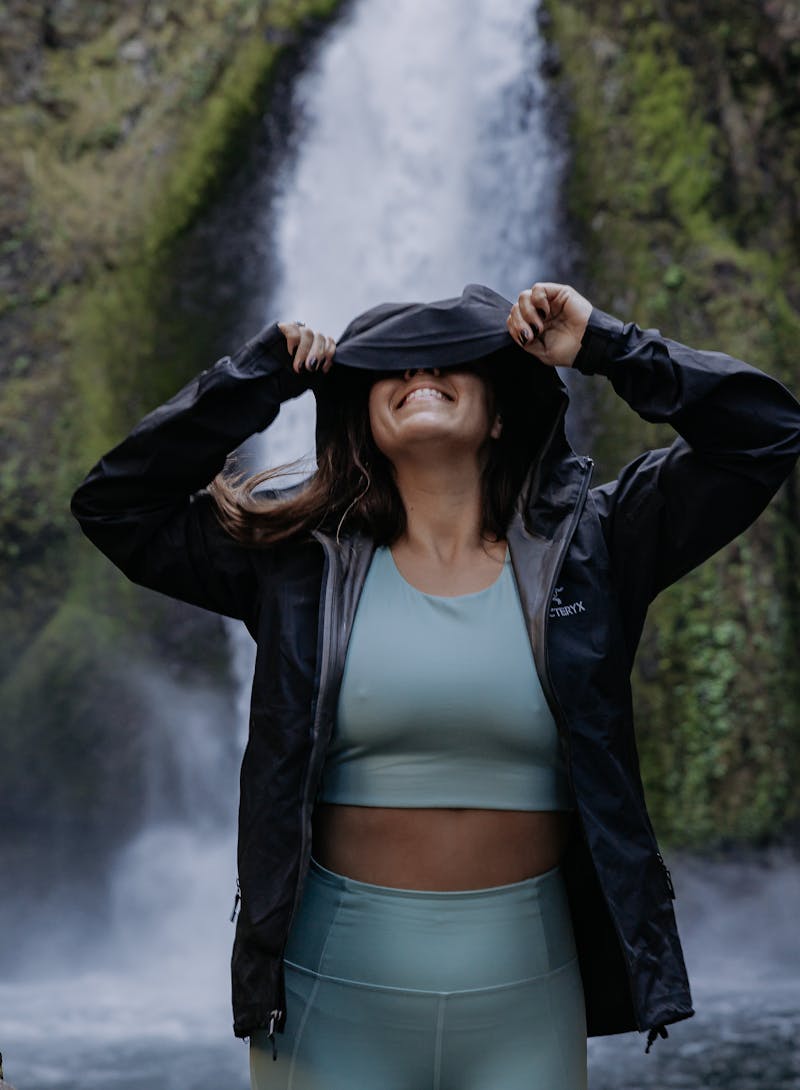 Lindsay Kagalis covering her head in the hood of a raincoat in front of a waterfall