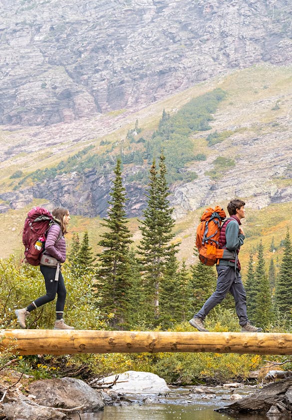 Two hikers walk over a wooden bridge over a mountain stream while backpacking in Oboz hiking boots.