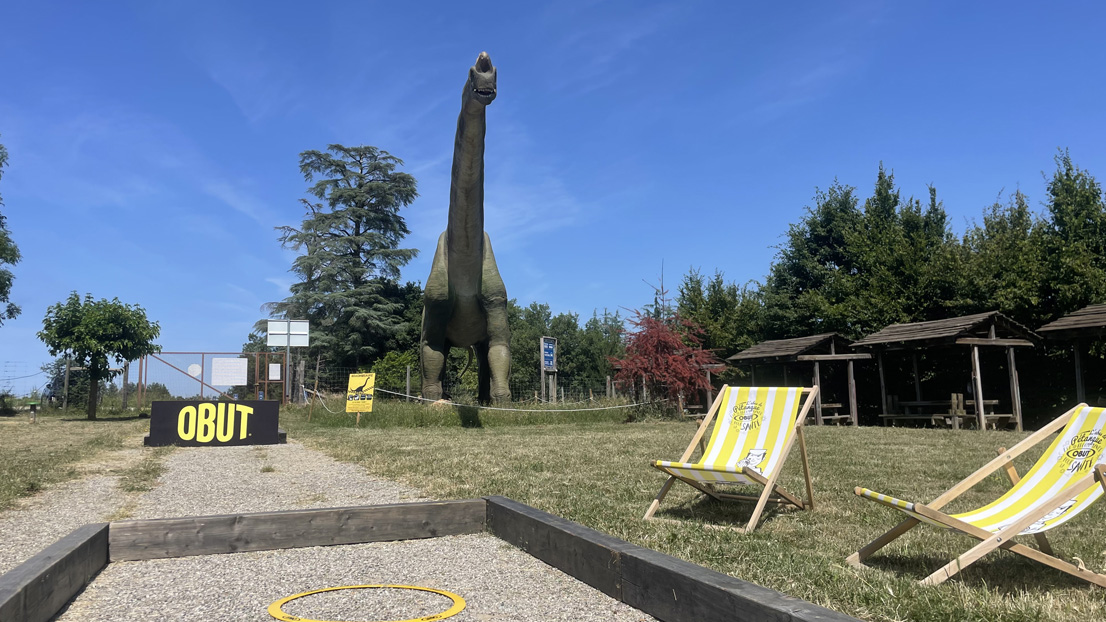 Vista de una pista de petanca Obut en el parque Wow Safari Peaugres, con tumbonas y un dinosaurio.