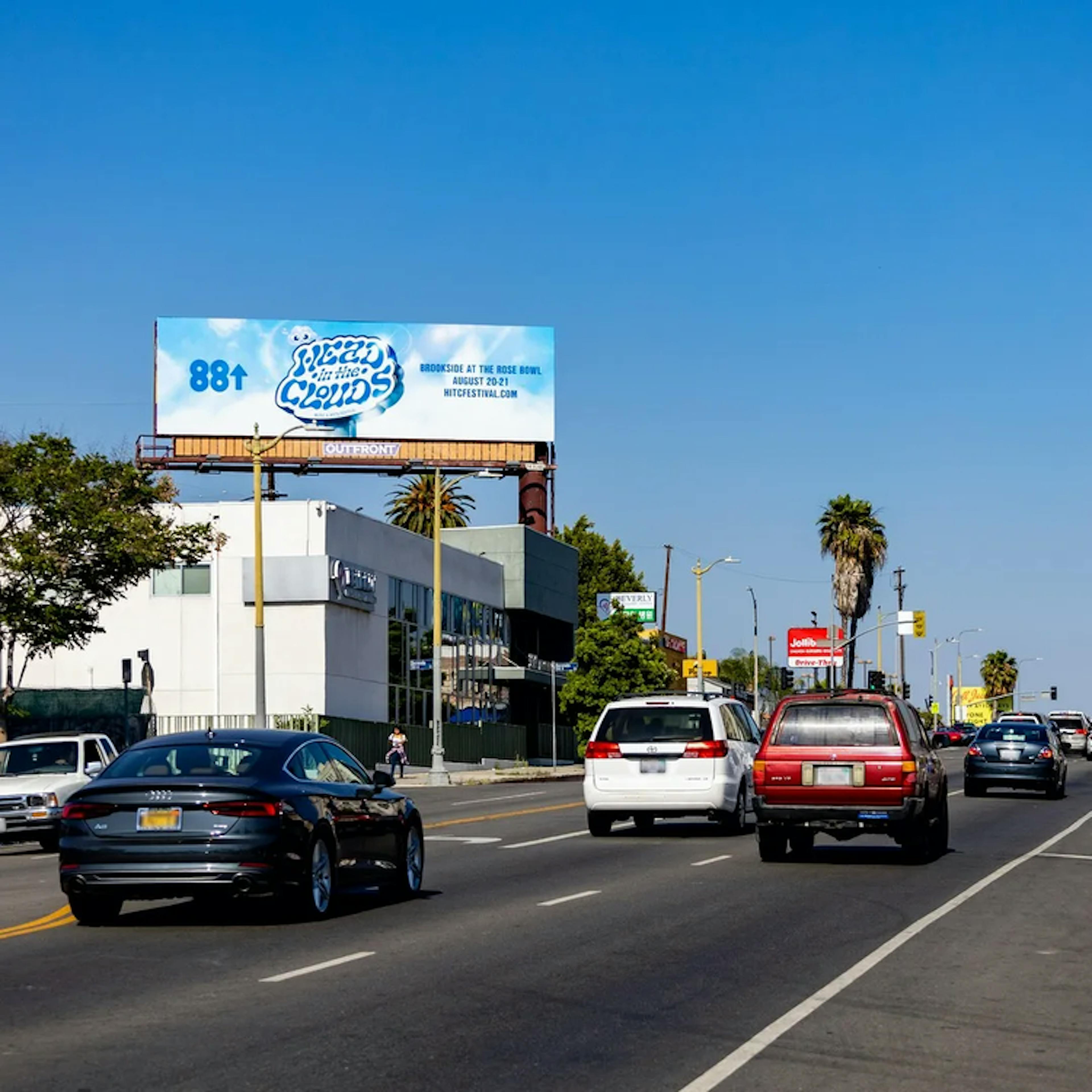 An image of a bustling city street under a clear blue sky, featuring a prominent billboard advertisement for the 'Head In The Clouds' Music & Arts Festival by 88rising. The billboard, set against the backdrop of a bright day, displays the festival's distinctive cloud-shaped logo in white and blue with the 88rising emblem, announcing the event at Brookside at the Rose Bowl on August 20-21. Vehicles are seen passing by on the road, indicating the billboard's roadside location in an active urban environment.