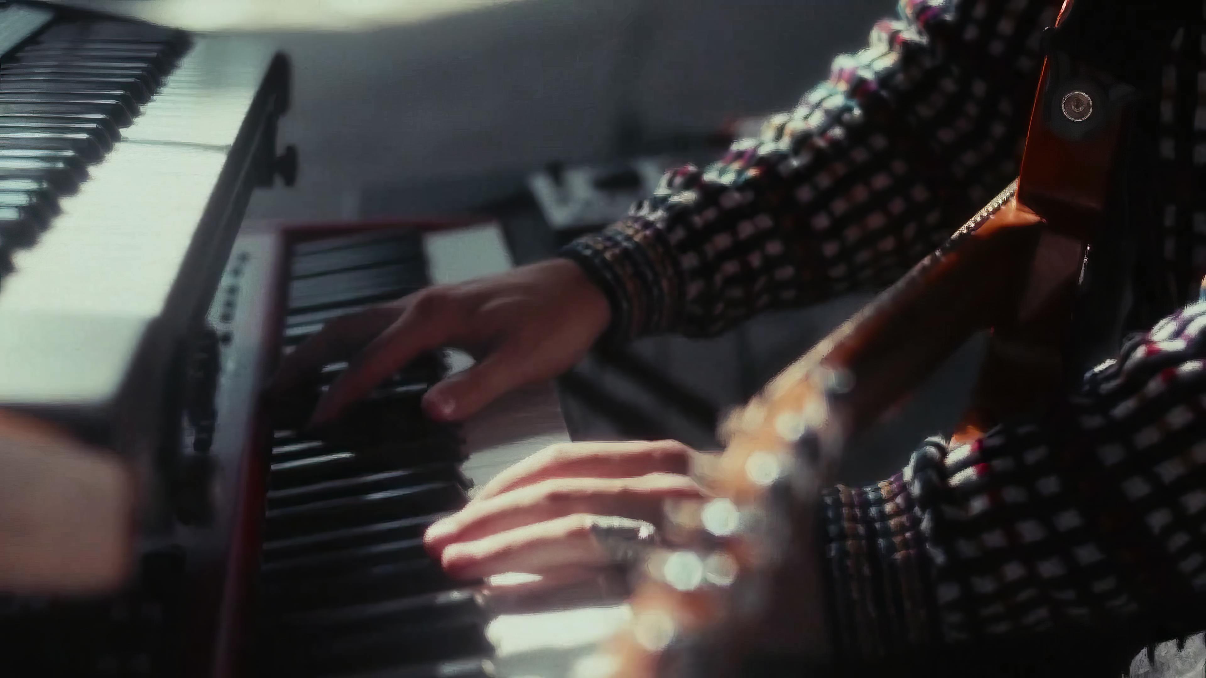 Ritt Momney is playing a keyboard, their hands pressing the keys. The musician is partially visible, dressed in a patterned top, with a glimpse of their face and headphones on. The scene is set indoors, and the lighting casts a warm, intimate glow on the instrument and the player.