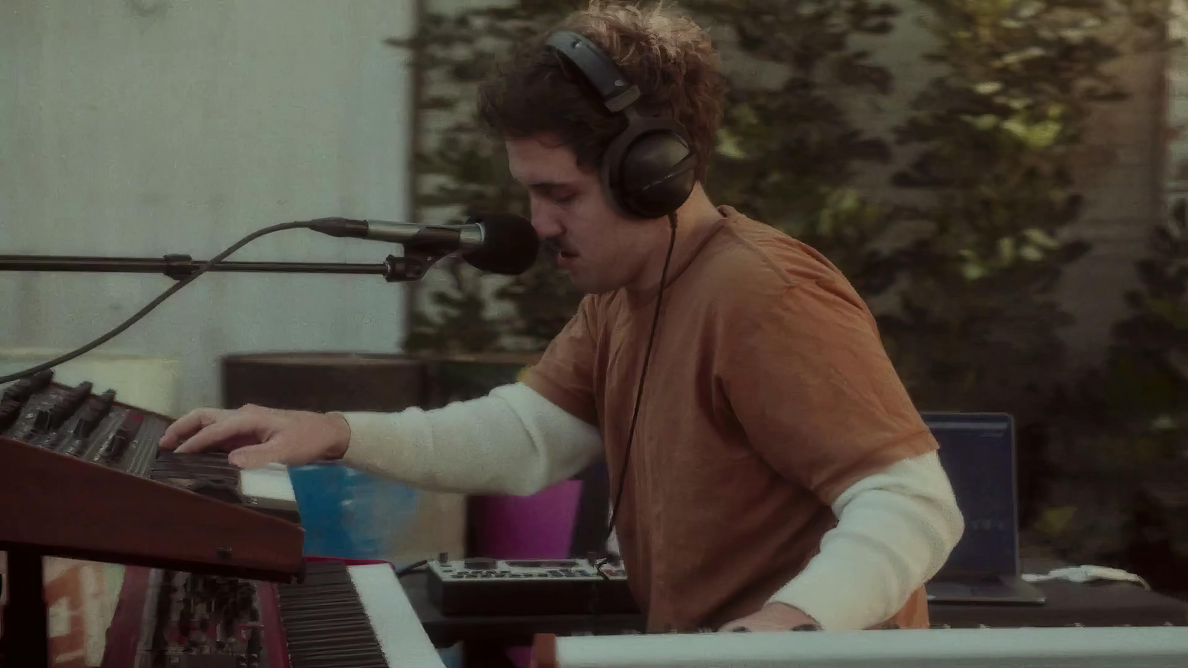Ritt Momney is performing on a keyboard for a live session. The musician, a young male with curly hair, is wearing a cream-colored sweater with rolled-up sleeves over an orange shirt. His focus is on the instrument, with a microphone set up in front of him, capturing the performance. The setting is outdoors with greenery in the background, suggesting a relaxed, natural atmosphere for the session.