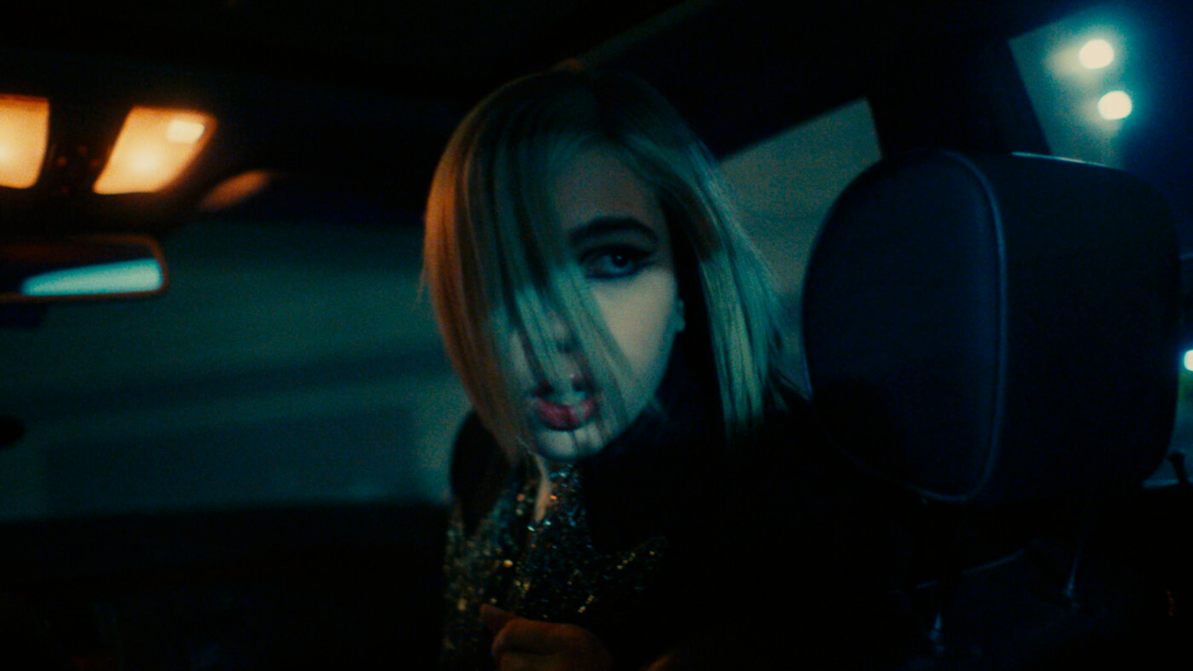 A woman with blonde hair and striking makeup looks intently through the car's front seats in a nighttime setting, in a still from Sunflower Bean's 'Who Put You Up To This?' music video.