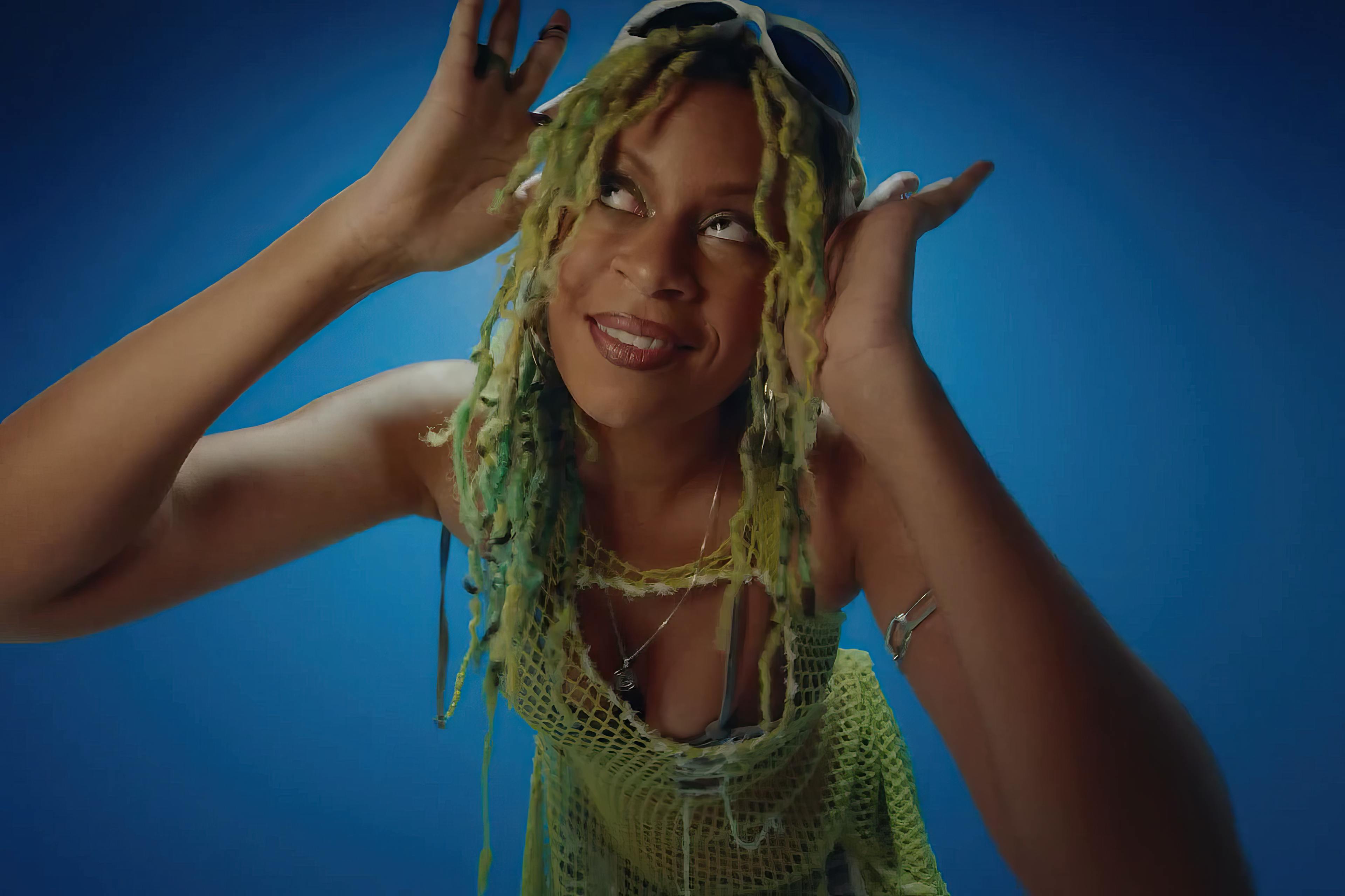 A joyful moment captured in the music video for 'Running Blind' by Aluna, Tchami, & Kareen Lomax. Aluna is seen with a beaming smile, her hands playfully adjusting her green and yellow tinted yarn-like hair. She's wearing a green crocheted top, adding a bright, summery vibe against a solid blue background that accentuates the cheerful and vibrant mood of the scene.