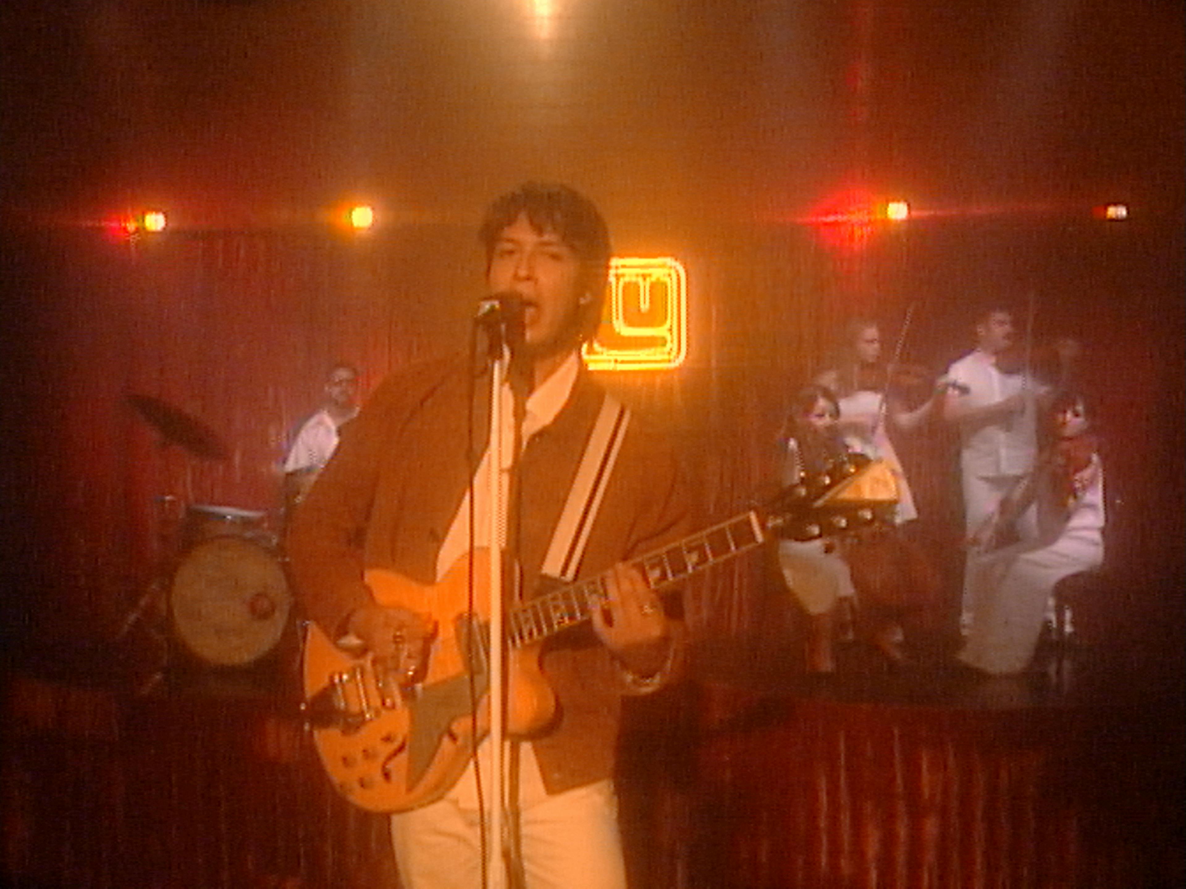 A vibrant scene from Inner Wave's music video 'Fever' features the band performing on stage. The focal point is a male singer with dark curly hair, holding a sunburst electric guitar, and singing into a microphone. He is wearing a brown jacket over a white shirt with a dark stripe. Behind him, the stage is bathed in a warm, reddish glow with a vintage feel.