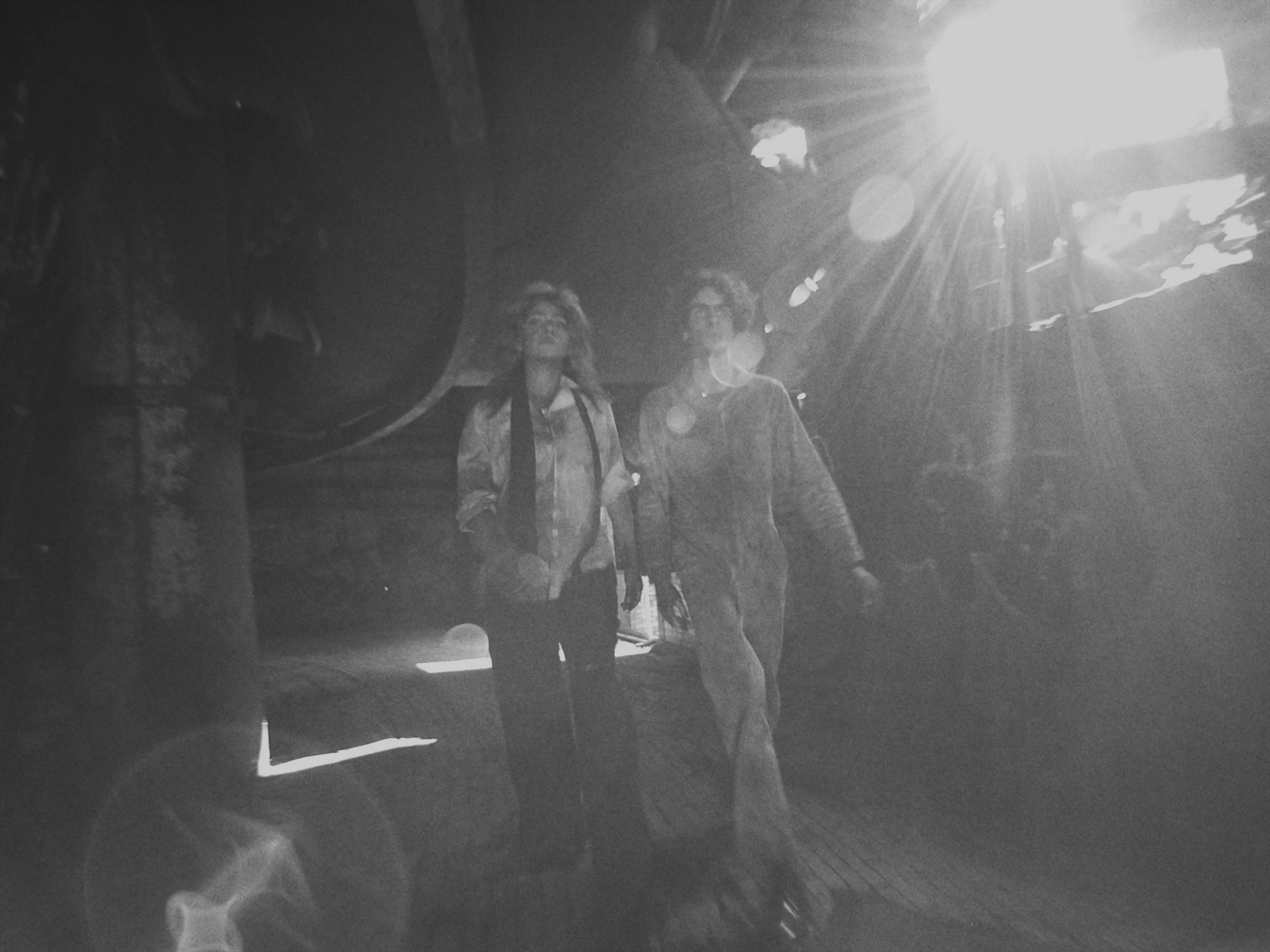 A black and white photo capturing two individuals walking through a beam of light in a dark, industrial setting. The light source, positioned at the top right corner, illuminates their figures, creating a dramatic contrast. Both individuals appear in mid-stride, one slightly ahead of the other, suggesting movement. The ambiance of the image is moody and cinematic, with the lens flare adding a raw, unfiltered quality to the moment captured.